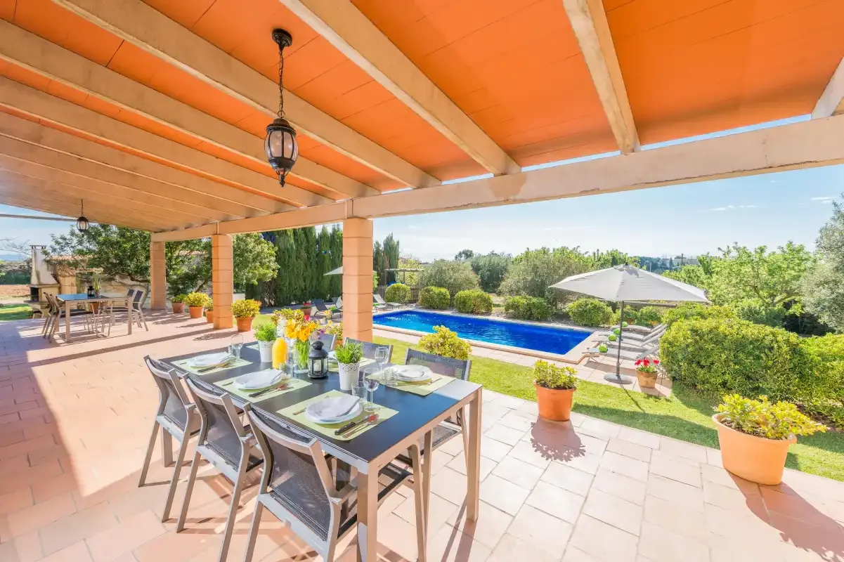 A covered patio with outdoor dining furniture overlooks a swimming pool and garden.