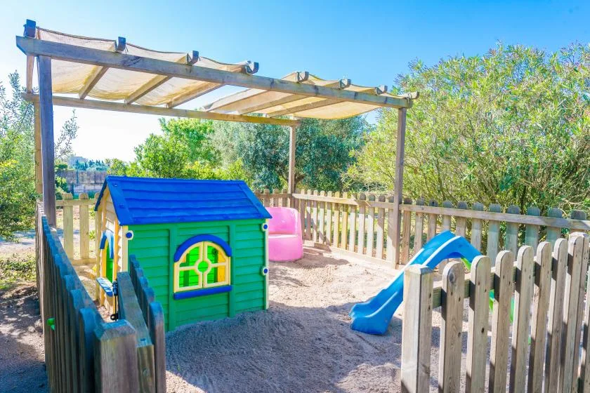 A fenced outdoor play area has a colorful playhouse and slide beneath a wooden canopy.