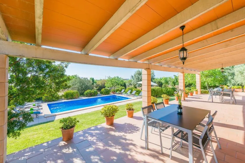 A covered patio with outdoor dining overlooks a swimming pool and garden.