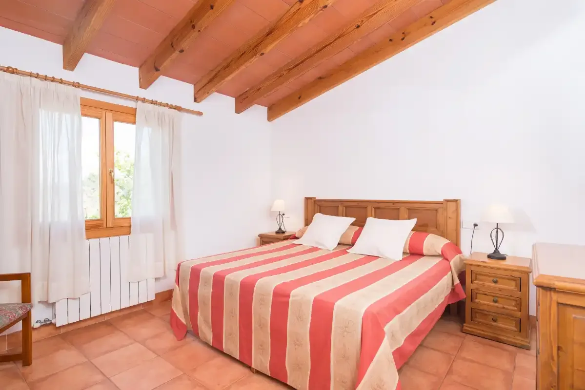 A bedroom with a striped bedspread, wooden ceiling beams, and a curtained window.