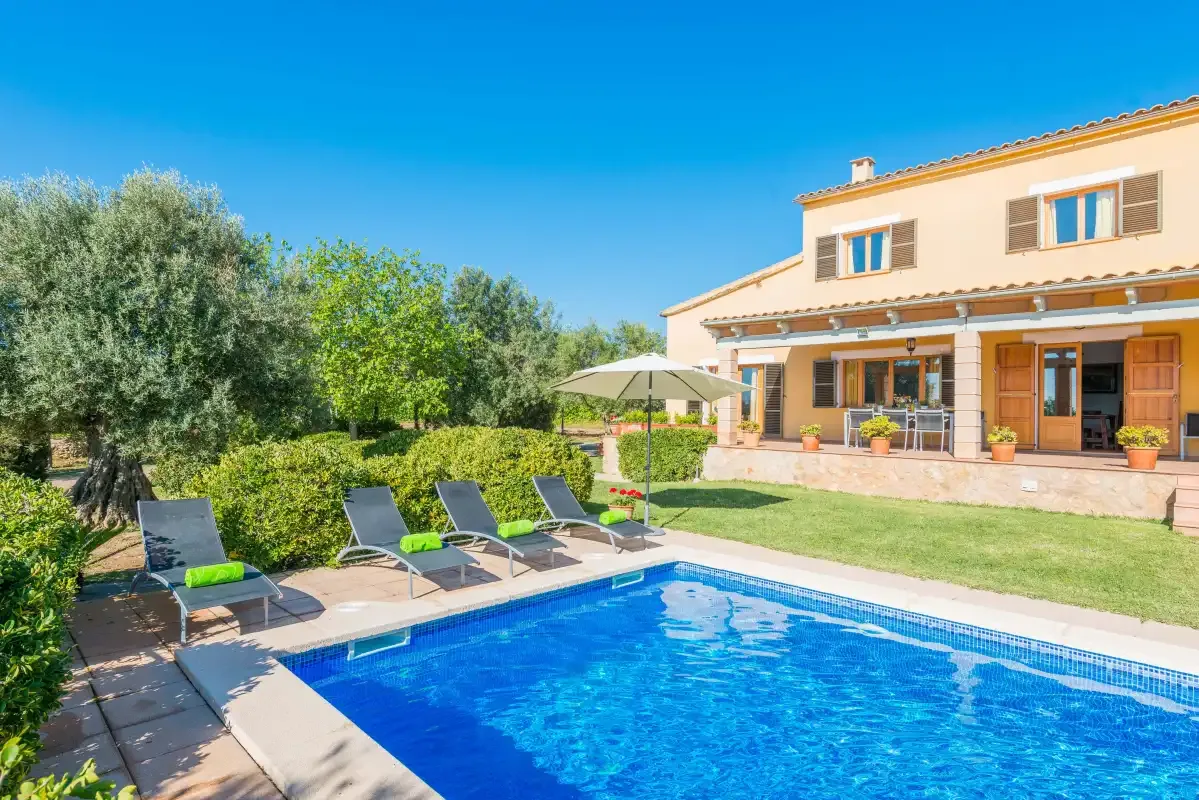 A swimming pool and lounge seating sit beside a house and green garden.