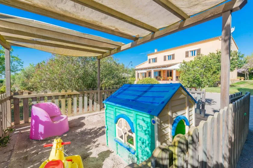 A fenced outdoor play area has a small playhouse and children’s toys beneath a canopy.