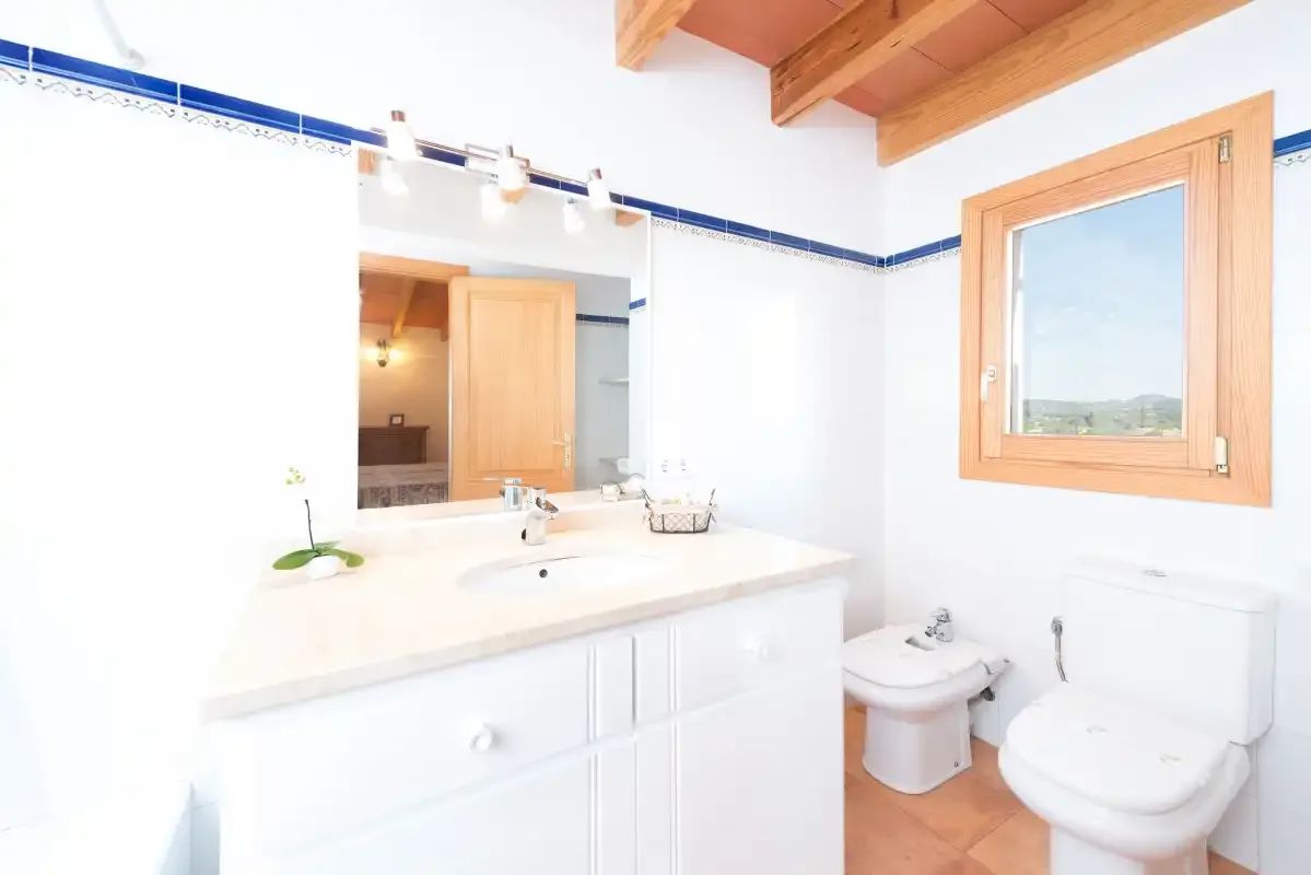 A bright bathroom has a vanity, toilet, and window beneath exposed wooden ceiling beams.