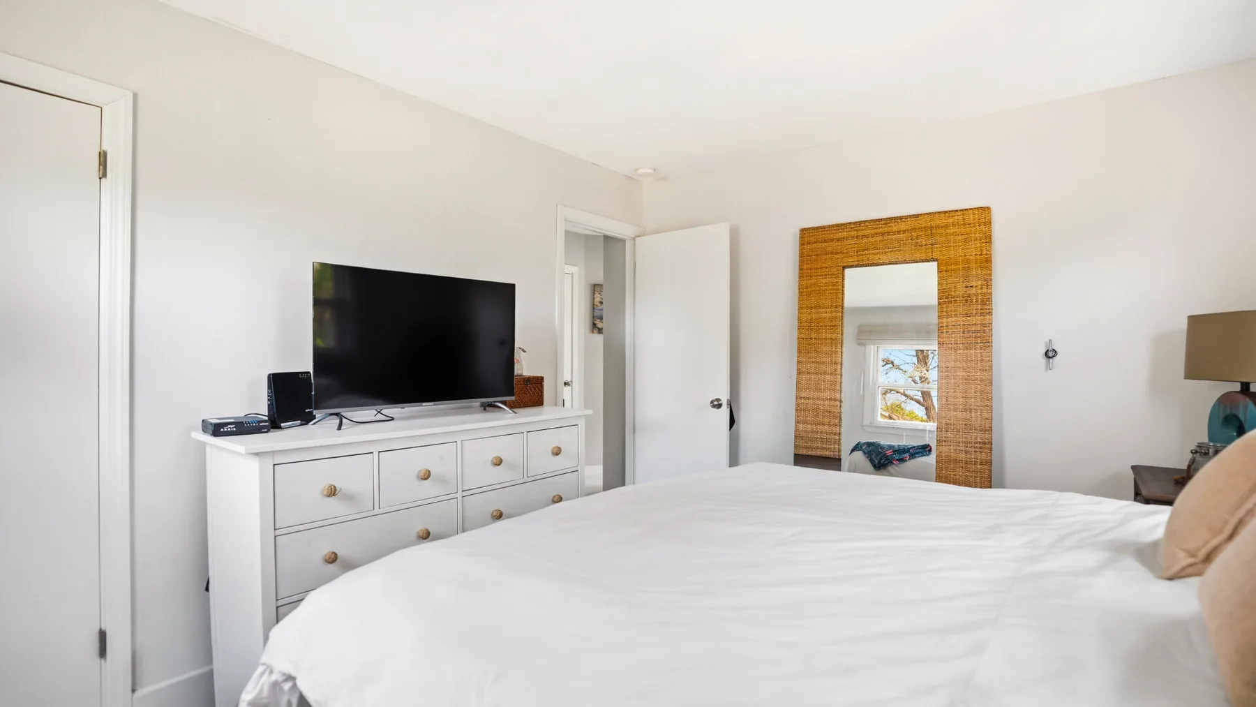 Un dormitorio luminoso con una cama blanca, una cómoda y un televisor.