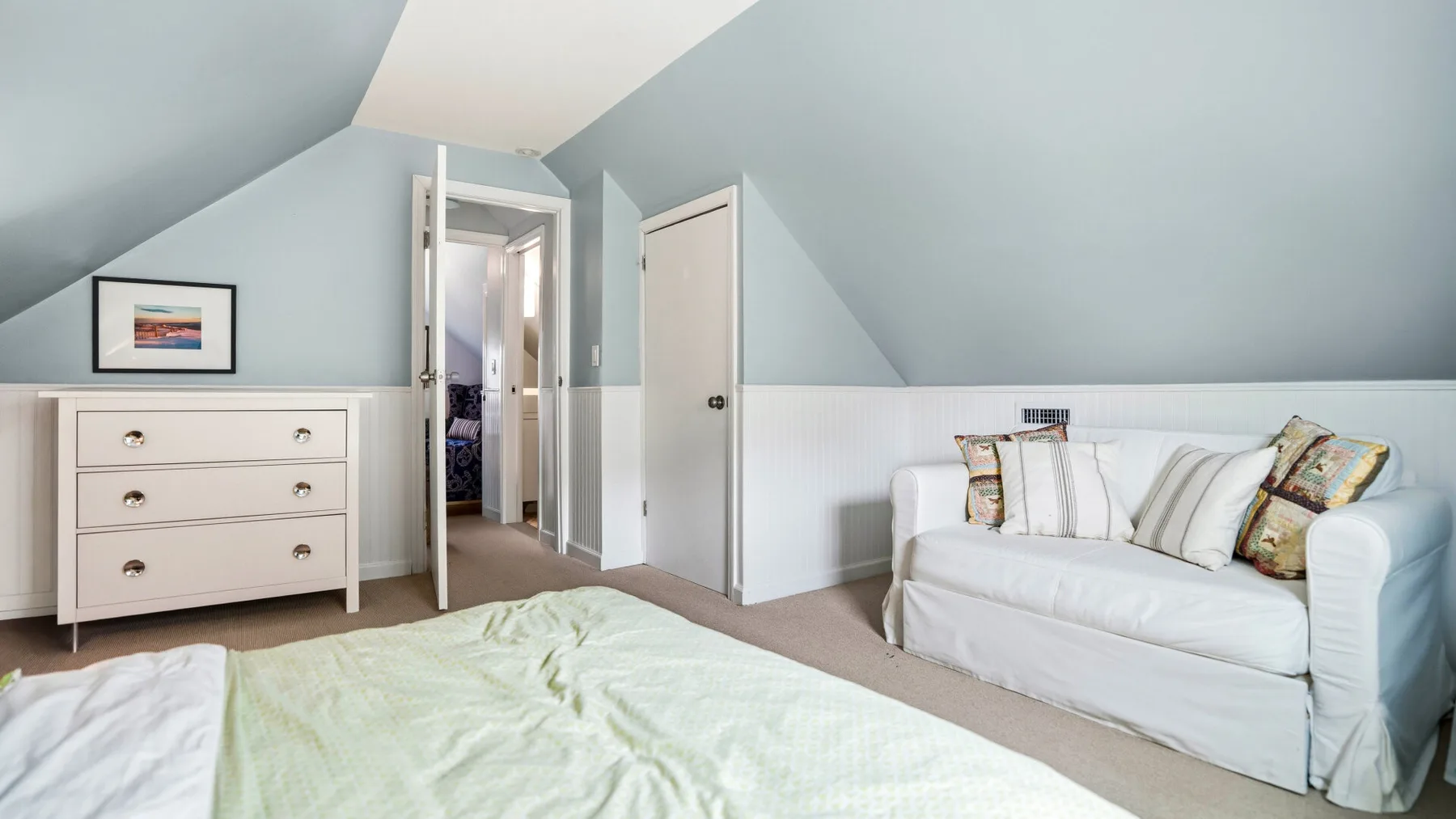 Un dormitorio luminoso con una cama y un sofá blanco bajo un techo inclinado.