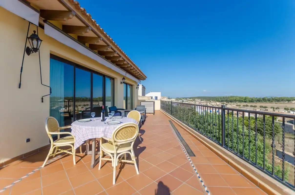 A sunny terrace has a dining table beside glass doors and a railing overlooking the landscape.
