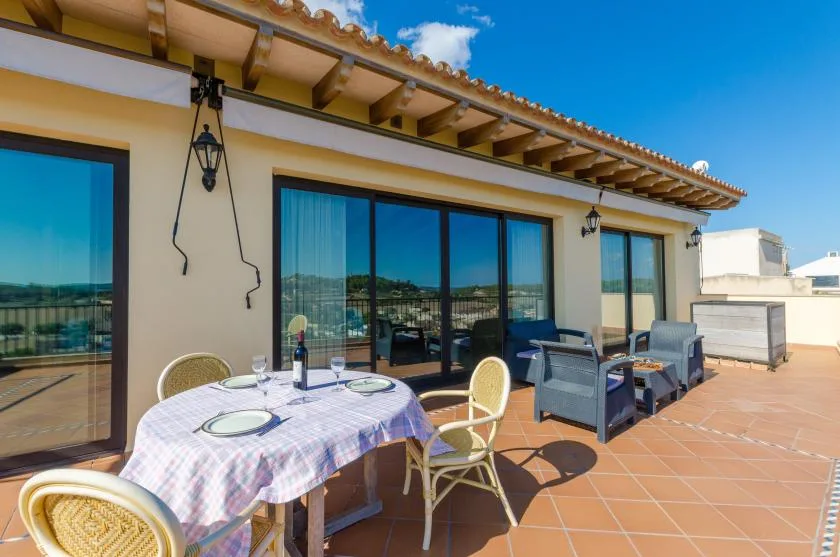A sunny outdoor terrace with a dining table, seating, and large glass doors.