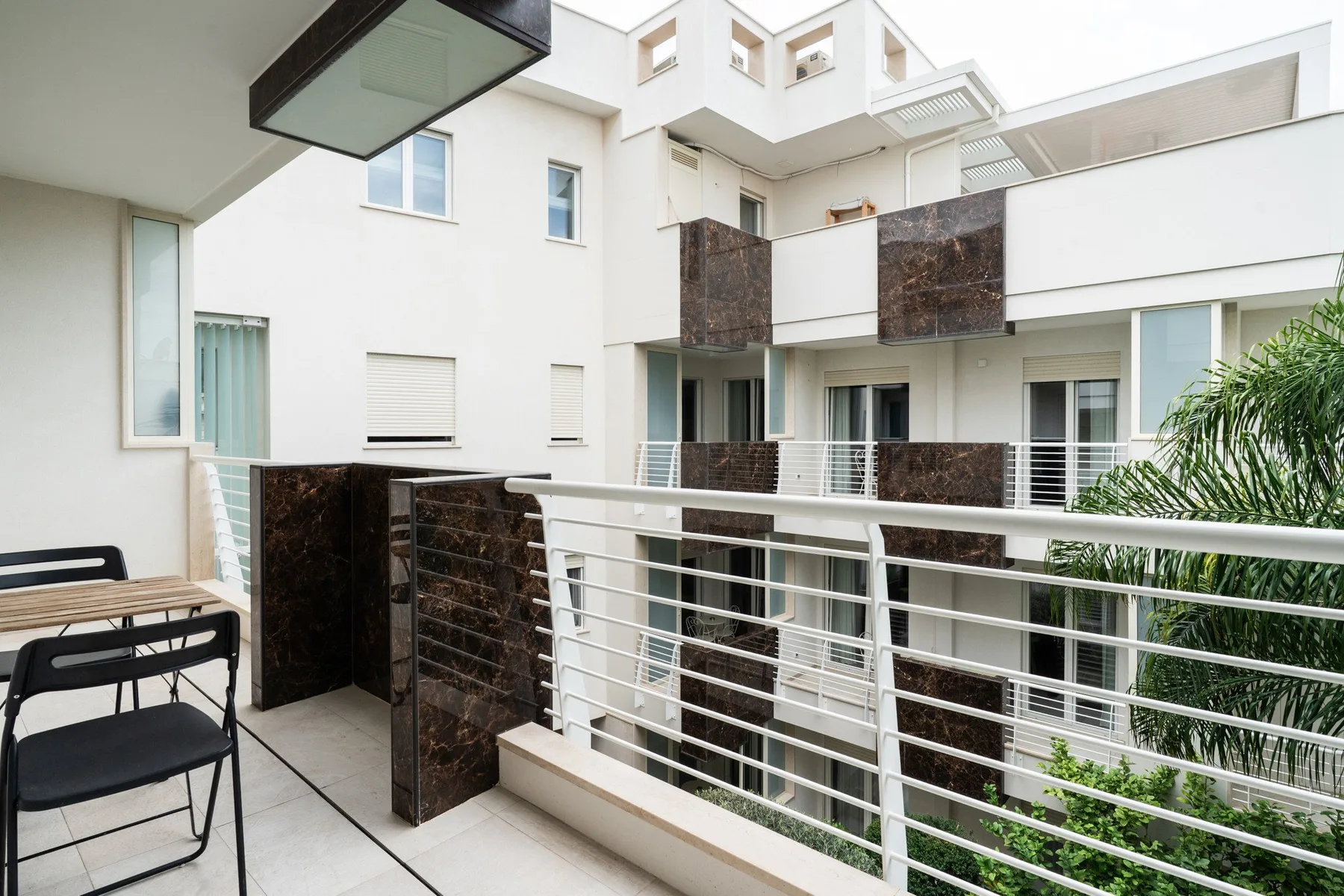 A modern outdoor balcony overlooks a white multi-story building, with metal railings and seating.