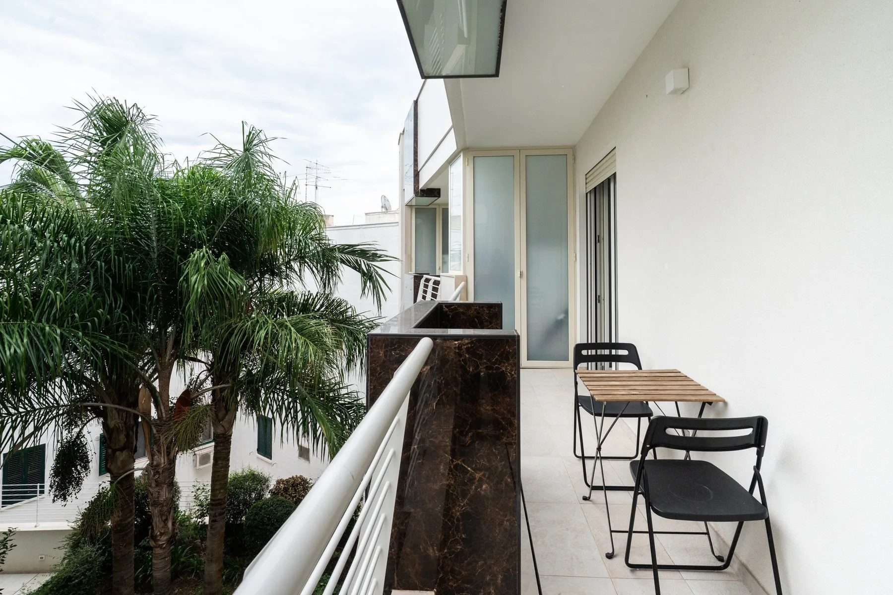 A narrow balcony has a small table and chairs beside a white wall.