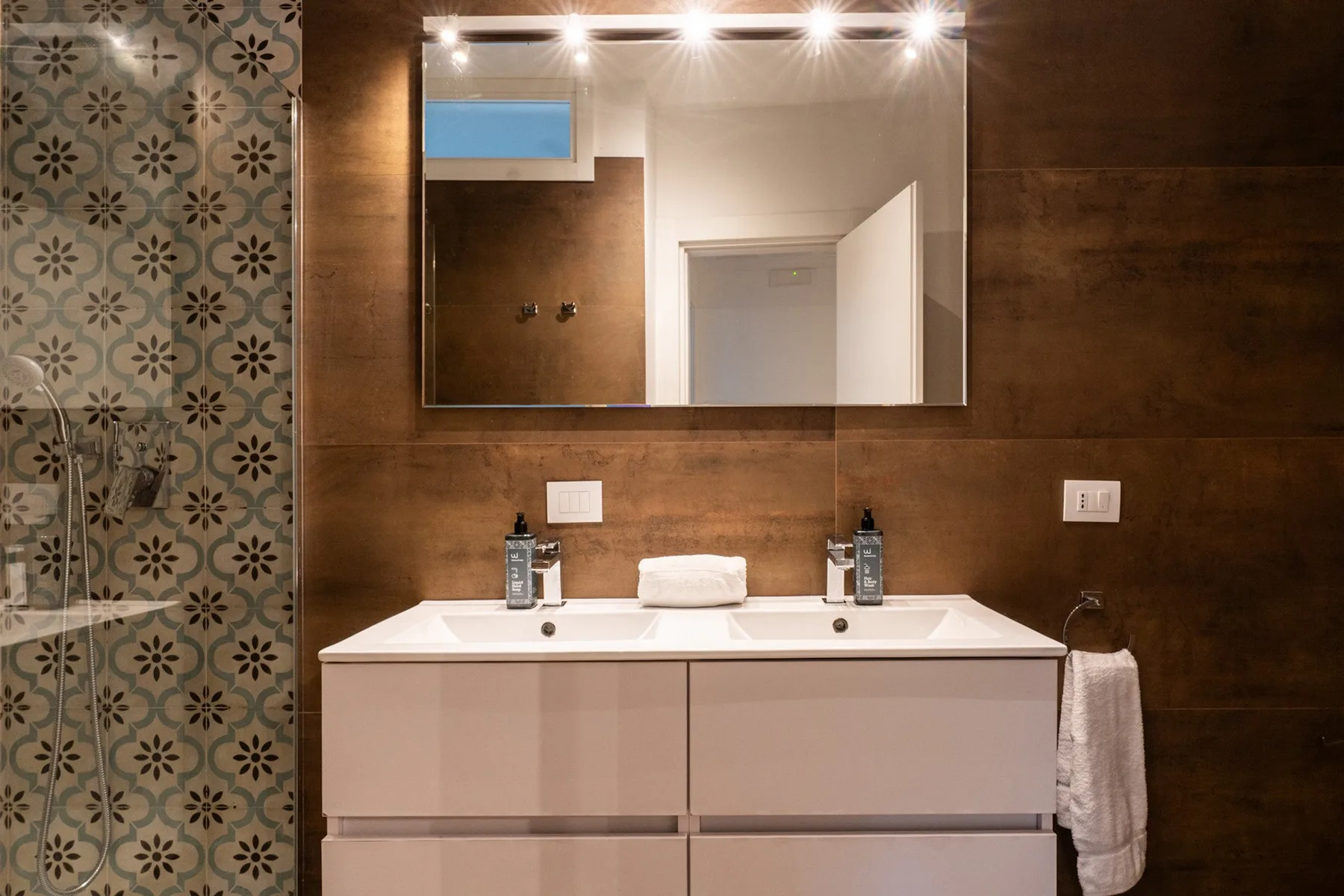 A bathroom vanity with a wide mirror and patterned tile along the shower.