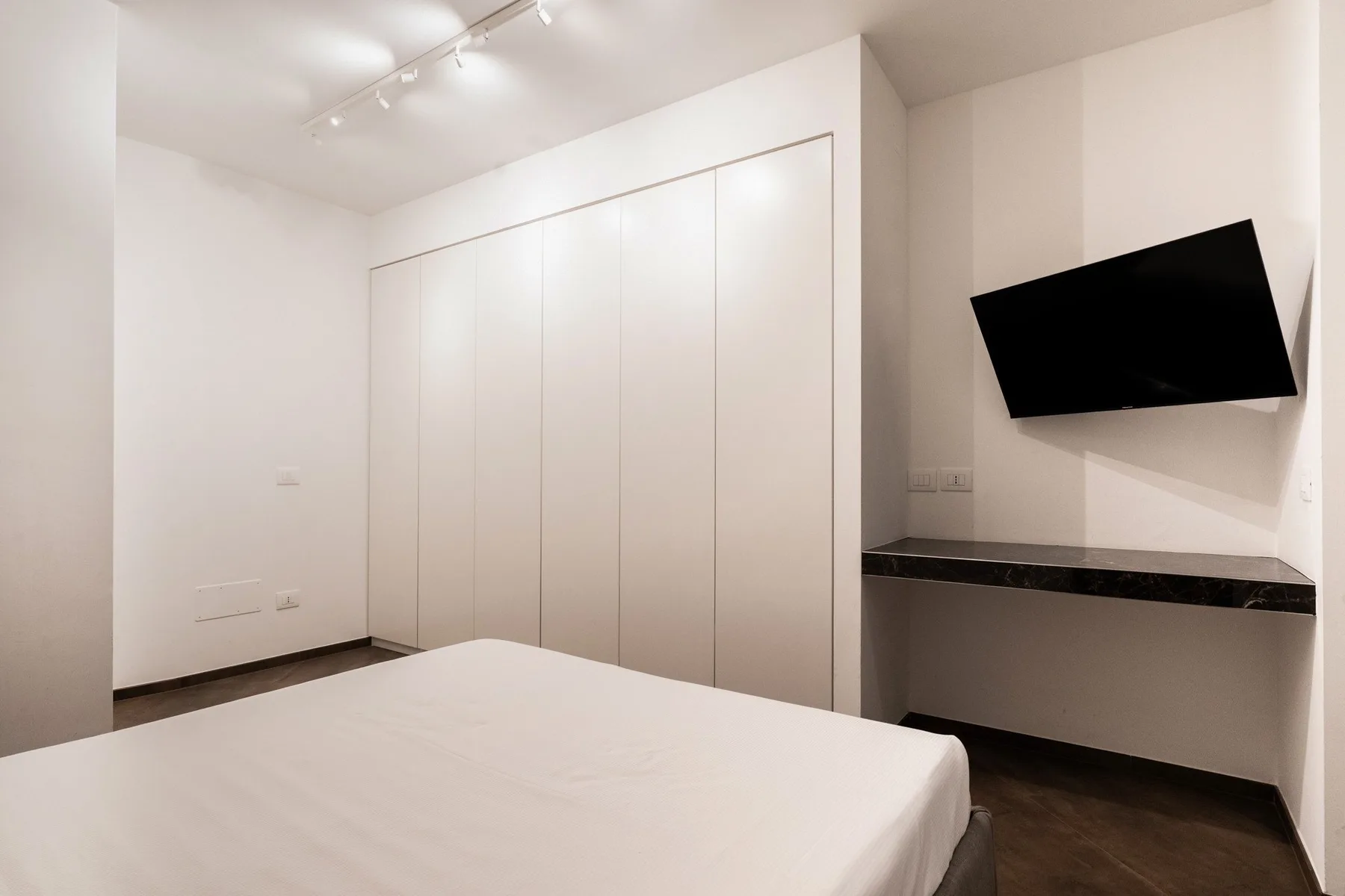 A bright bedroom with a white bed, built-in cabinets, and a wall-mounted television.