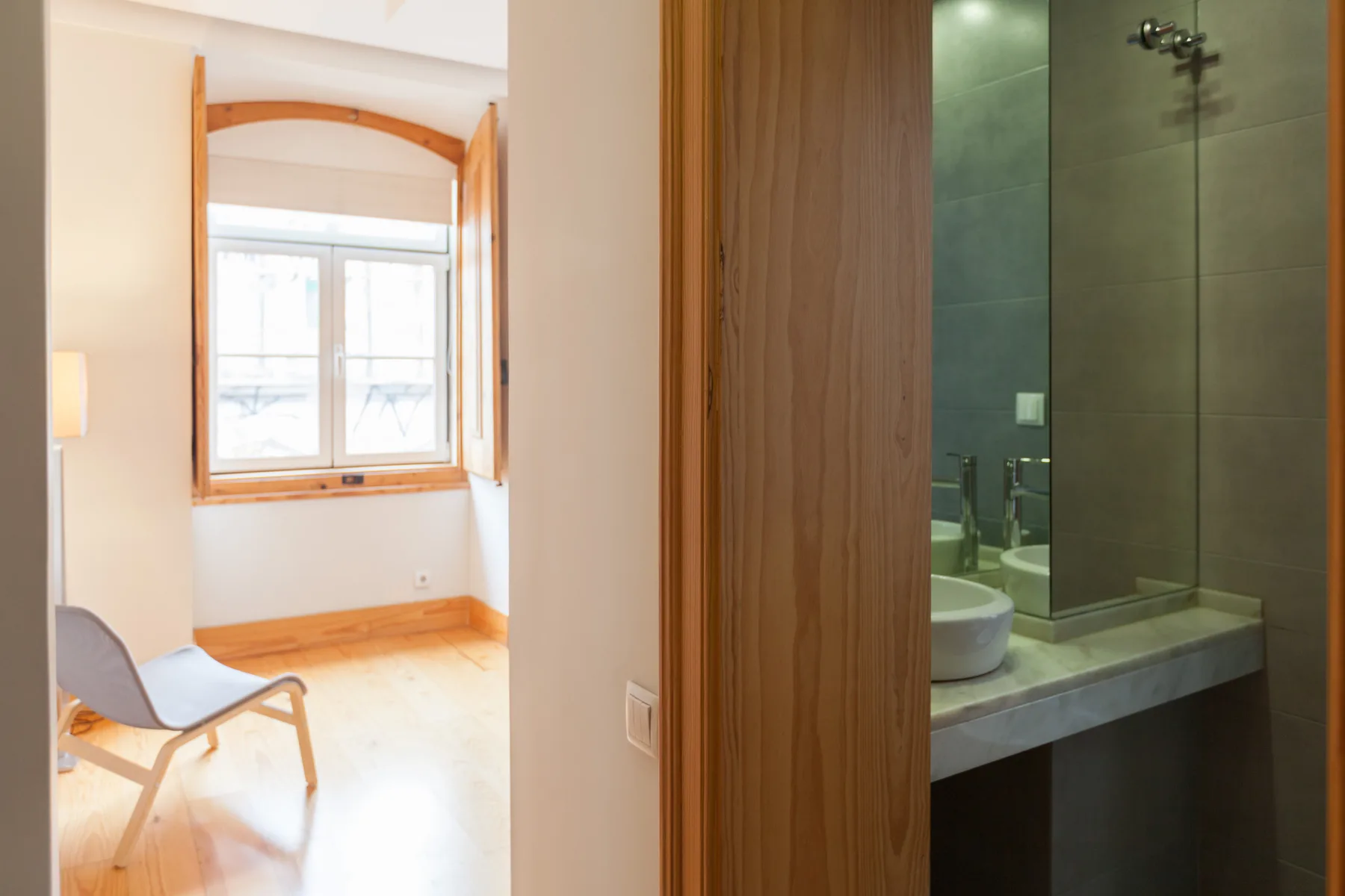 Una habitación luminosa con ventana y silla da a un baño con ducha.
