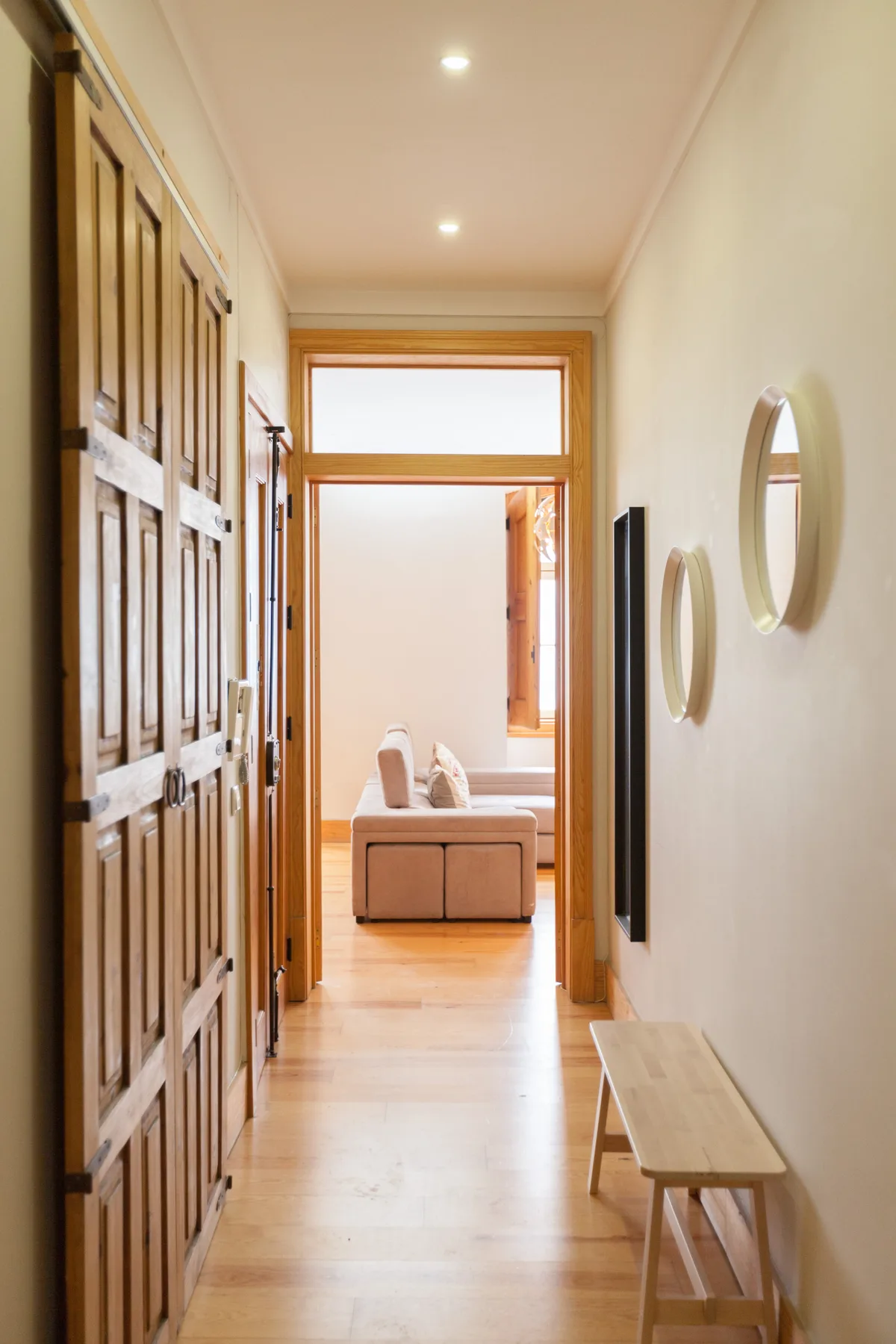 Un pasillo luminoso con puertas de madera, un banco y vistas a una zona de estar.