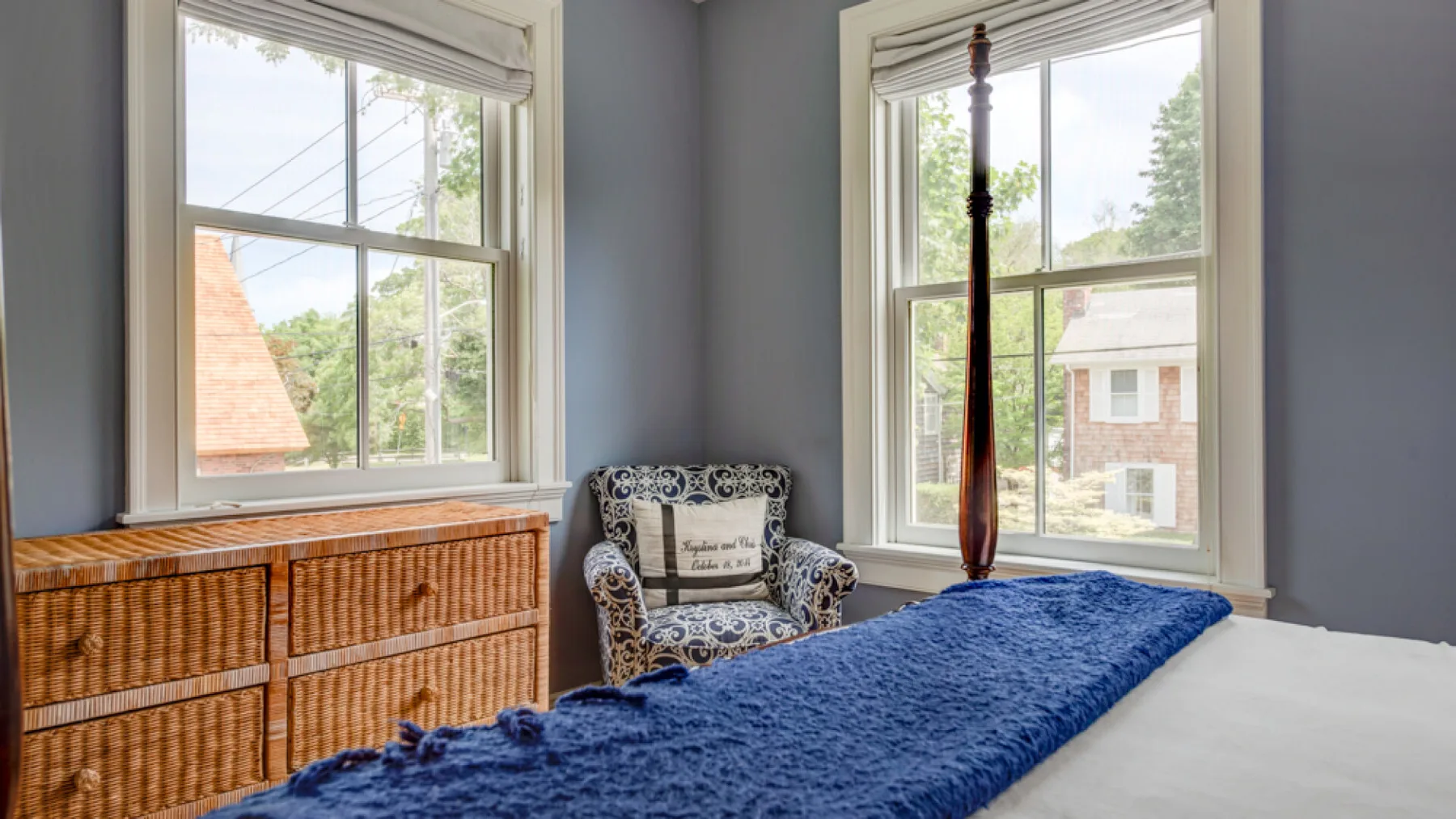 A bedroom with a blue bedspread, an armchair, and two large windows.
