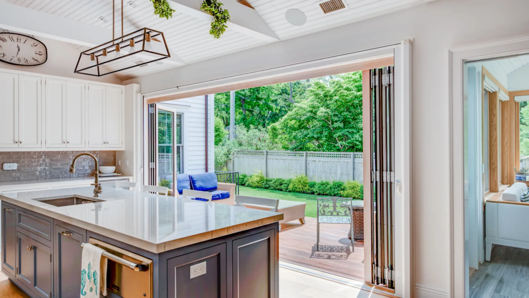 A bright kitchen opens onto a deck with outdoor seating and greenery.