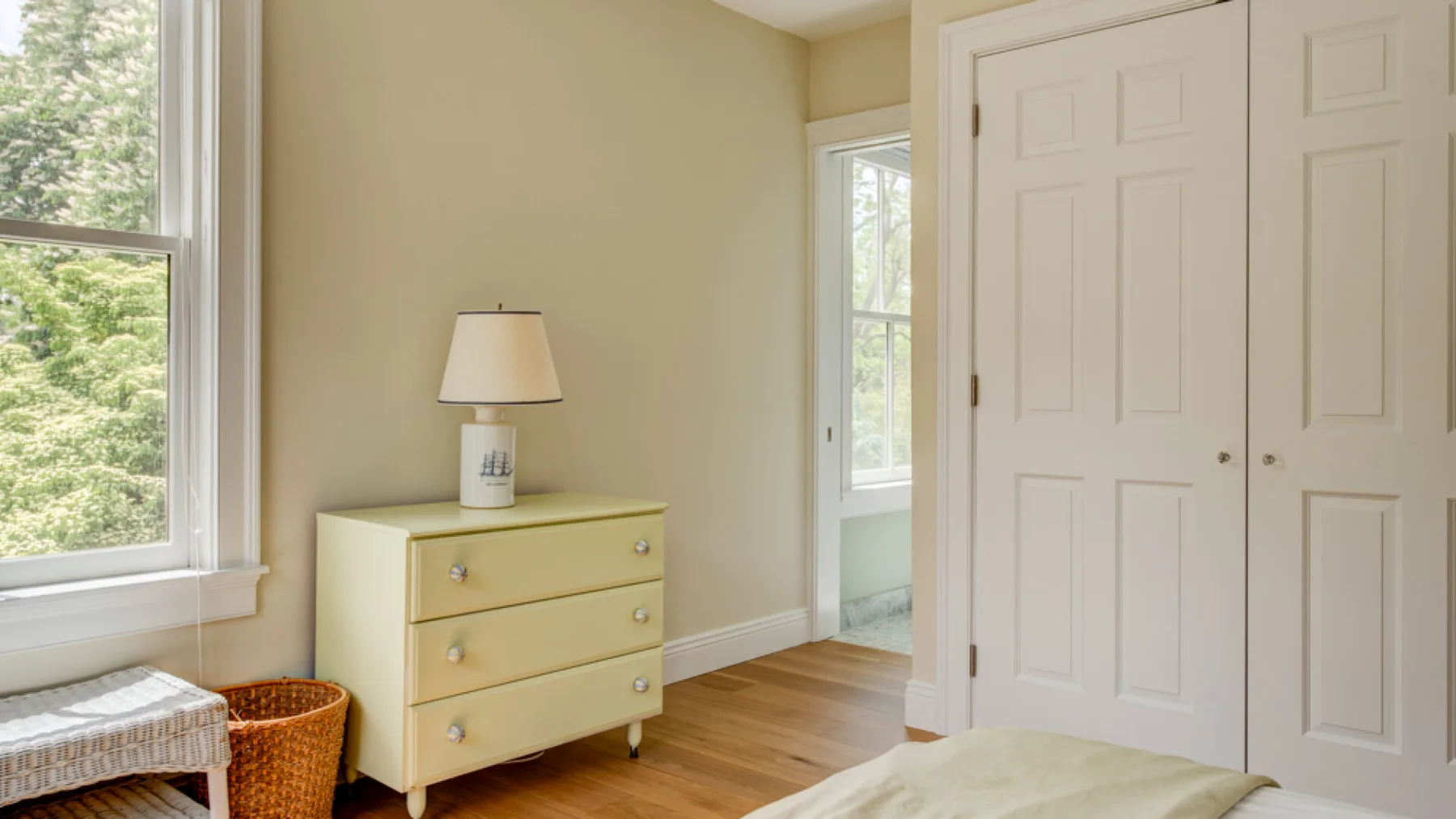 A bright bedroom with a pale dresser, large windows, and wood flooring.