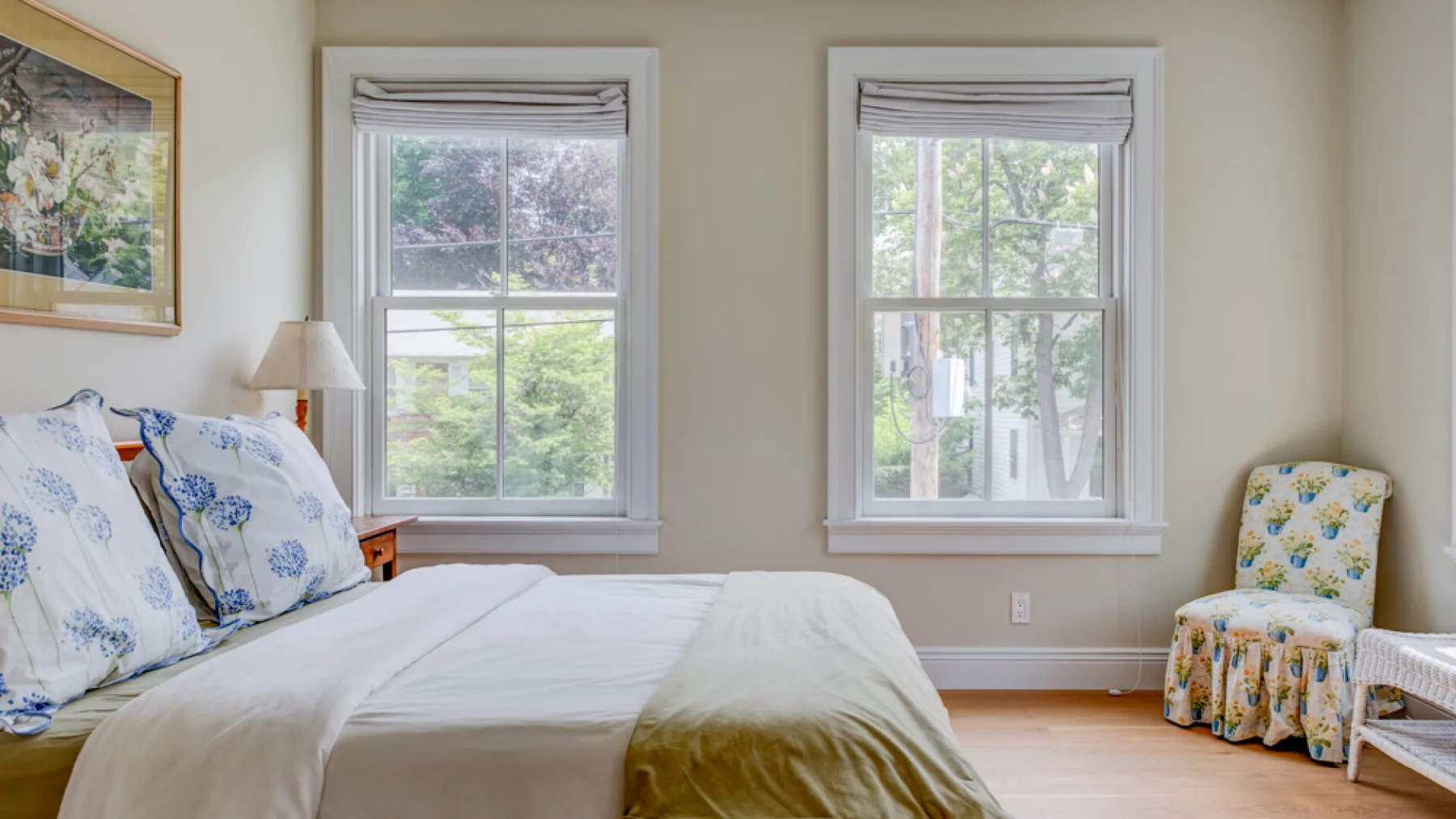A bedroom with a bed and two windows overlooking leafy trees.