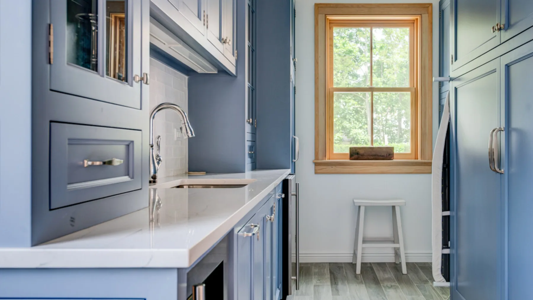 A blue kitchen with white countertops and a window above a small seating area.
