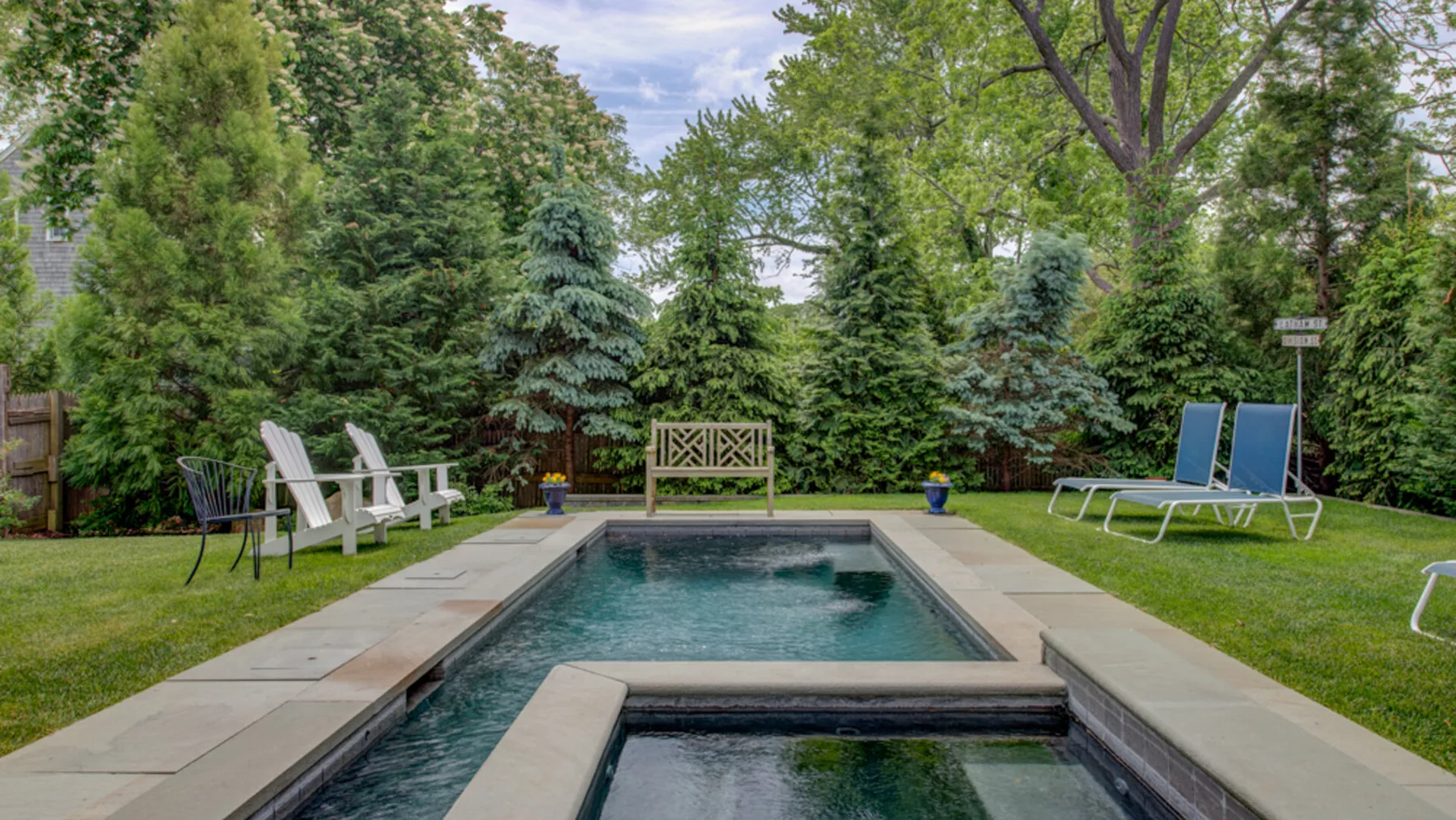 An outdoor swimming pool is surrounded by lawn, trees, and lounge seating.