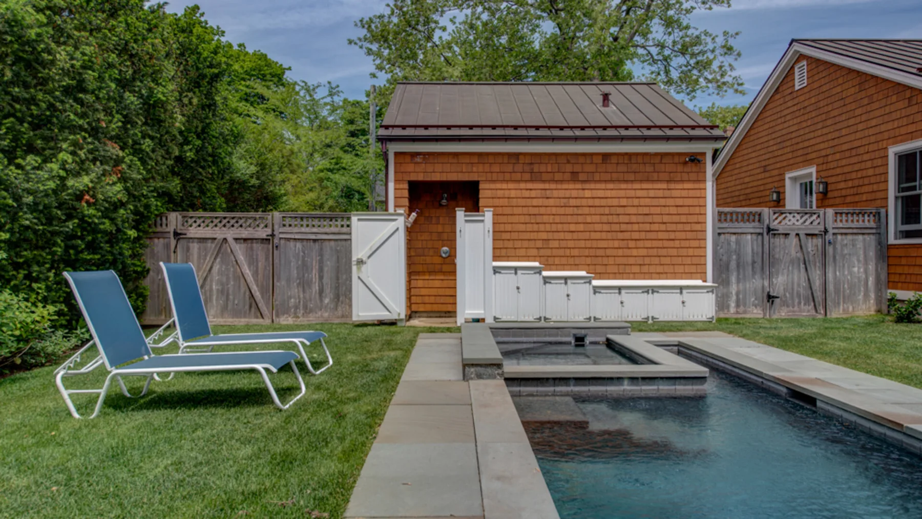 An outdoor swimming pool sits beside a wooden building and fenced yard.