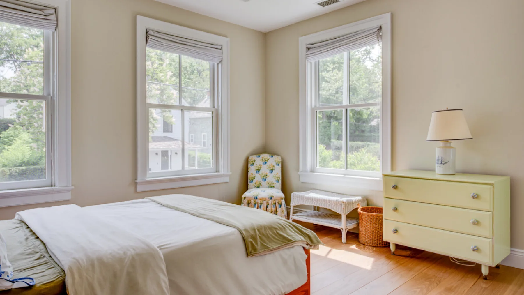 A bright bedroom with a bed, pale dresser, and windows overlooking greenery.