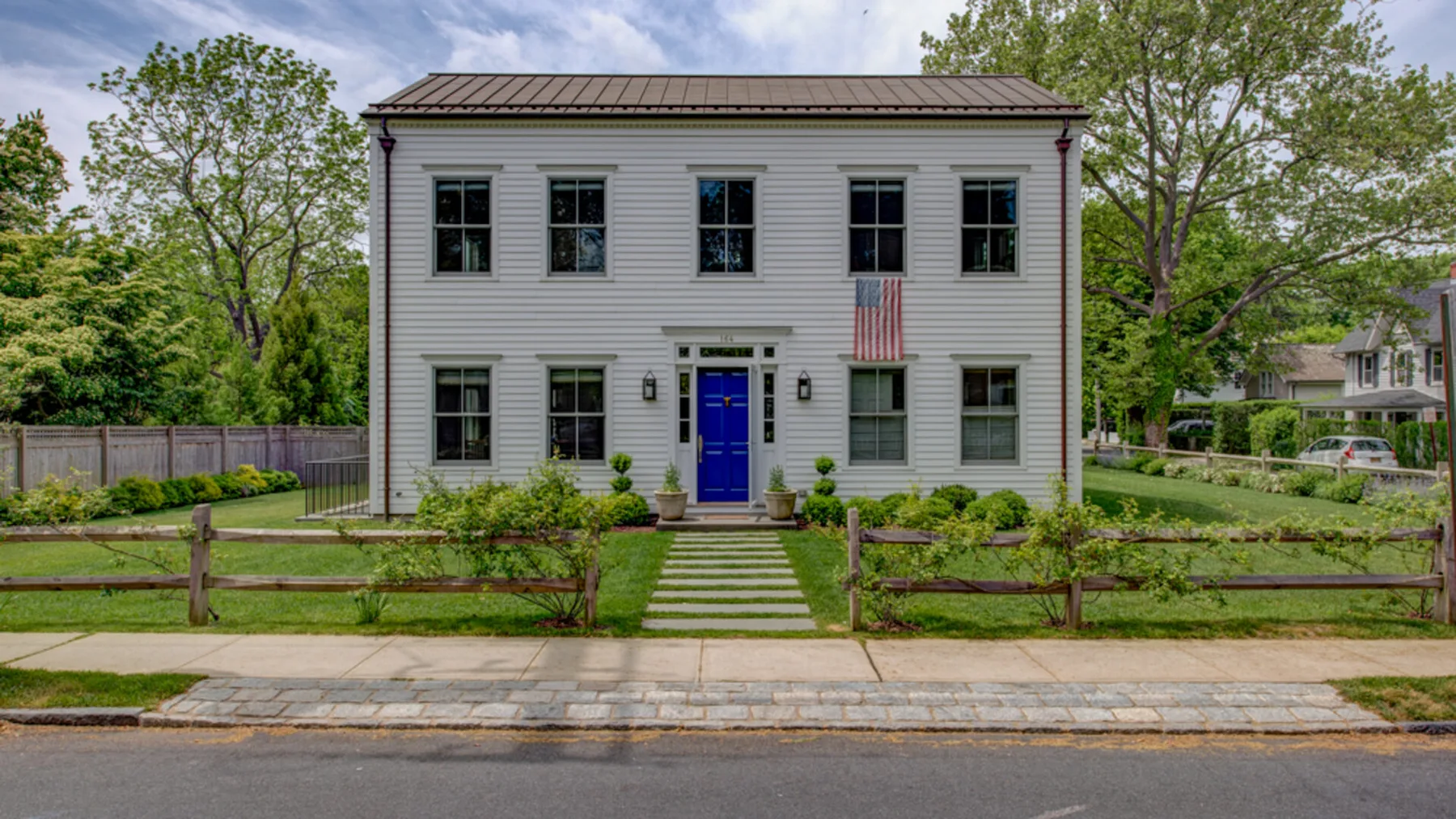 A two-story house stands behind a low wooden fence, with a path leading to its blue front door.