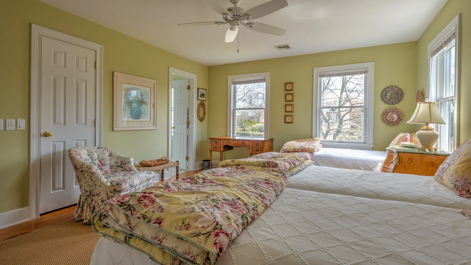 Un dormitorio con dos camas, ropa de cama floral y ventanas con vistas a los árboles.