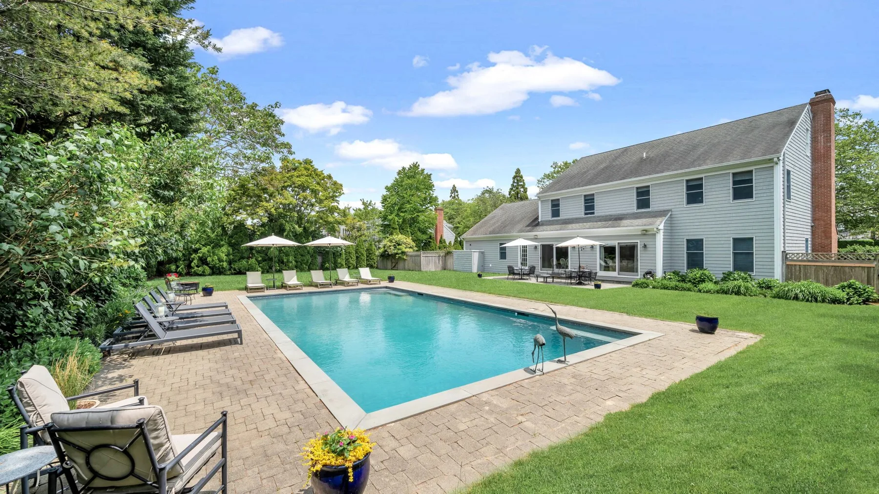 Una piscina en el patio junto a una casa grande, con pavimento y asientos.
