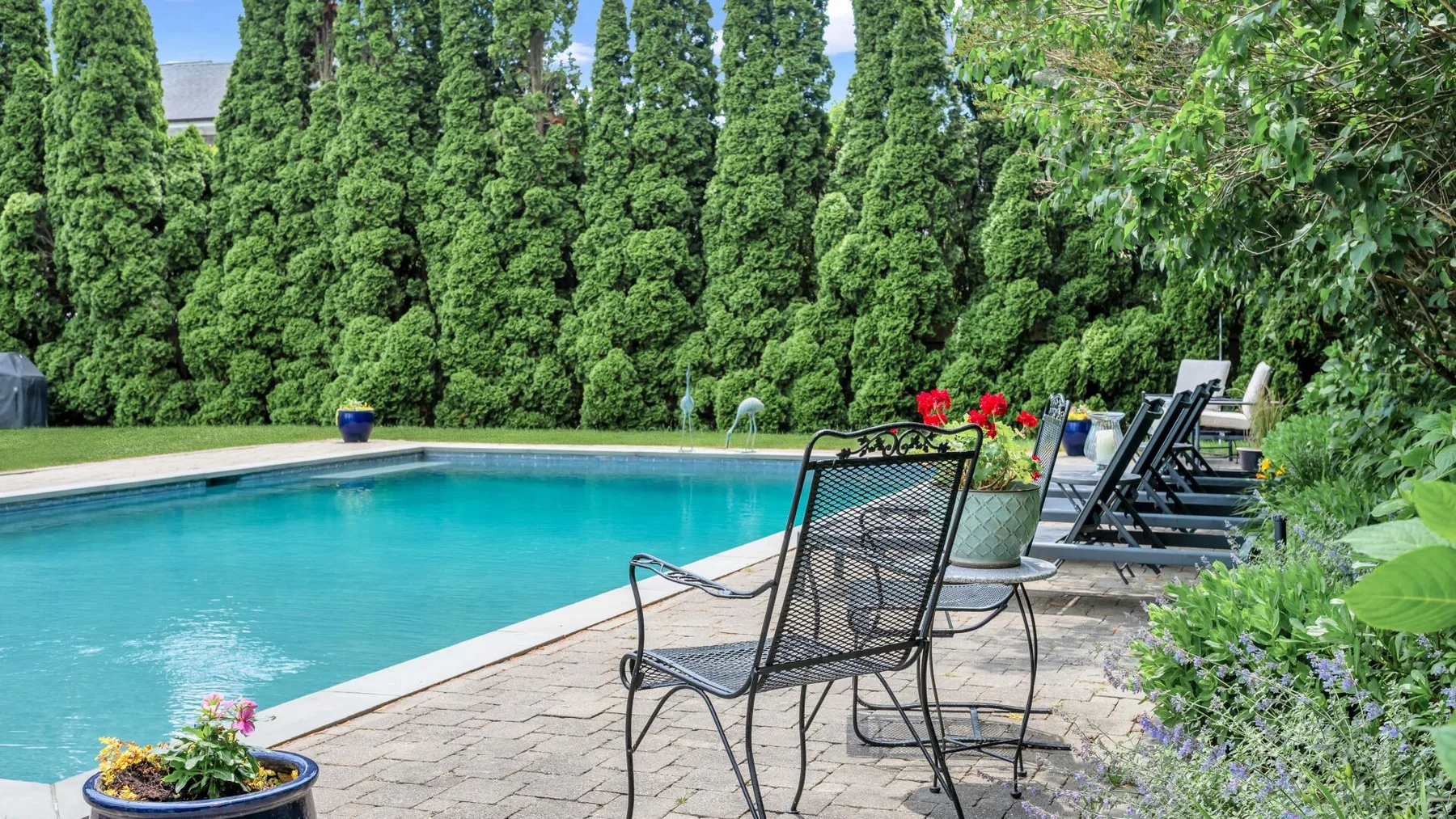 Una piscina al aire libre junto a un patio pavimentado con asientos y vegetación.