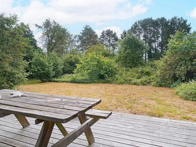 Una terraza de madera da a un patio de hierba seca rodeado de árboles y arbustos.