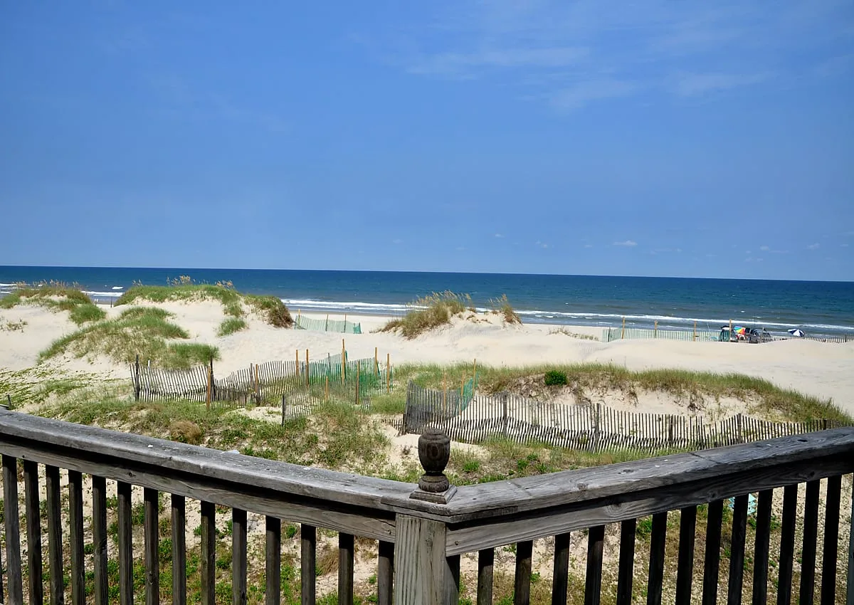 A wooden deck railing overlooks sandy dunes and the ocean.