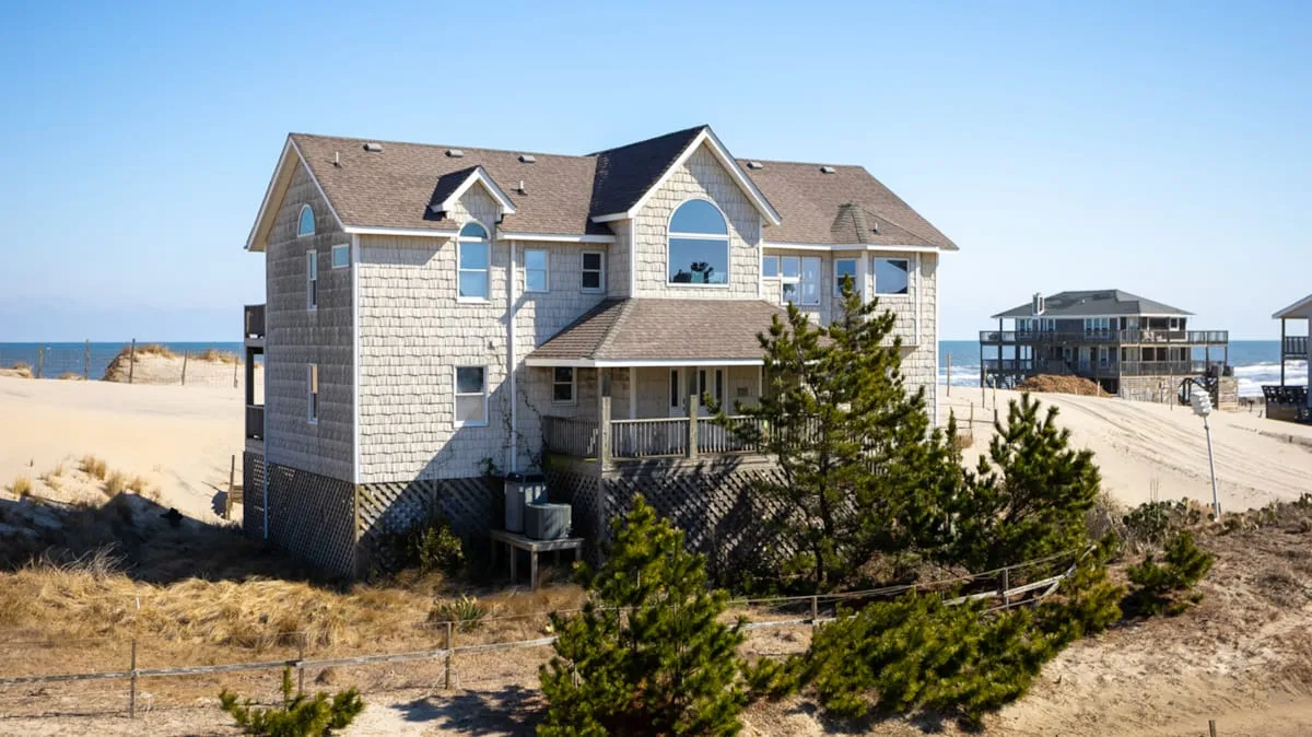 A large coastal house stands among sandy dunes beside the ocean.