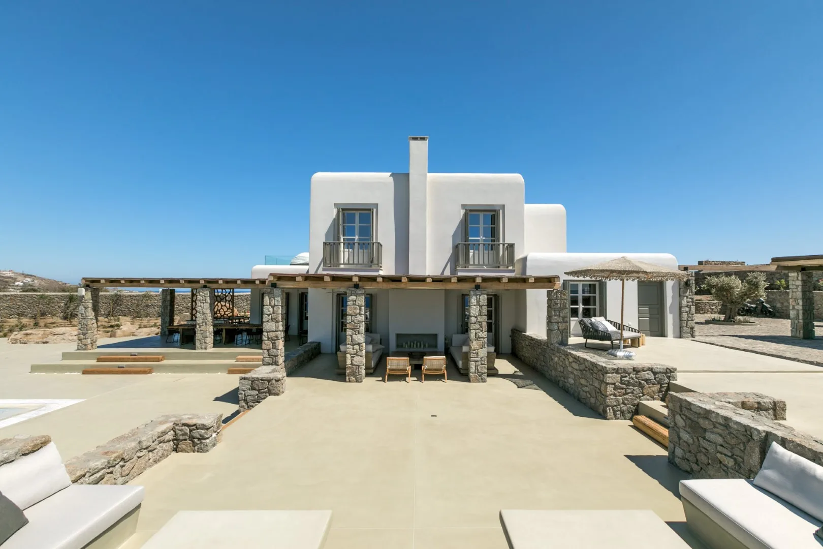 A white two-story villa opens onto a broad stone terrace beneath a clear blue sky.