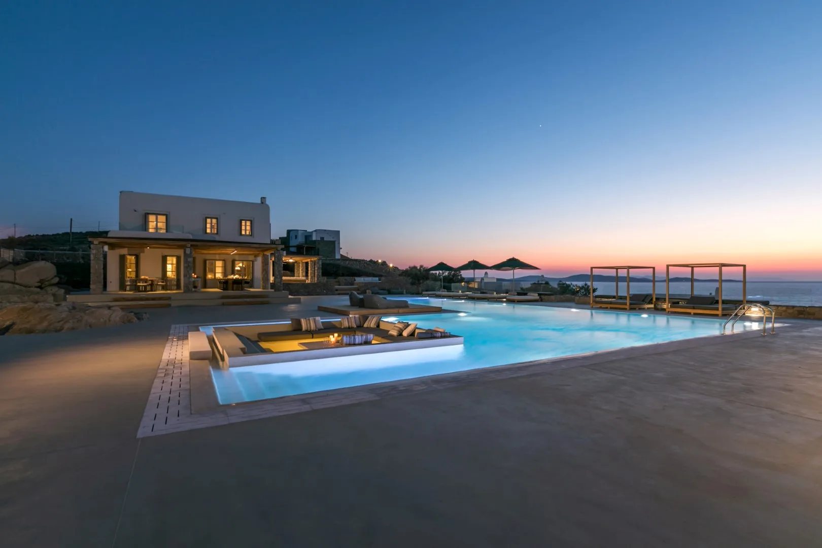 An outdoor swimming pool glows beside a modern house at dusk.