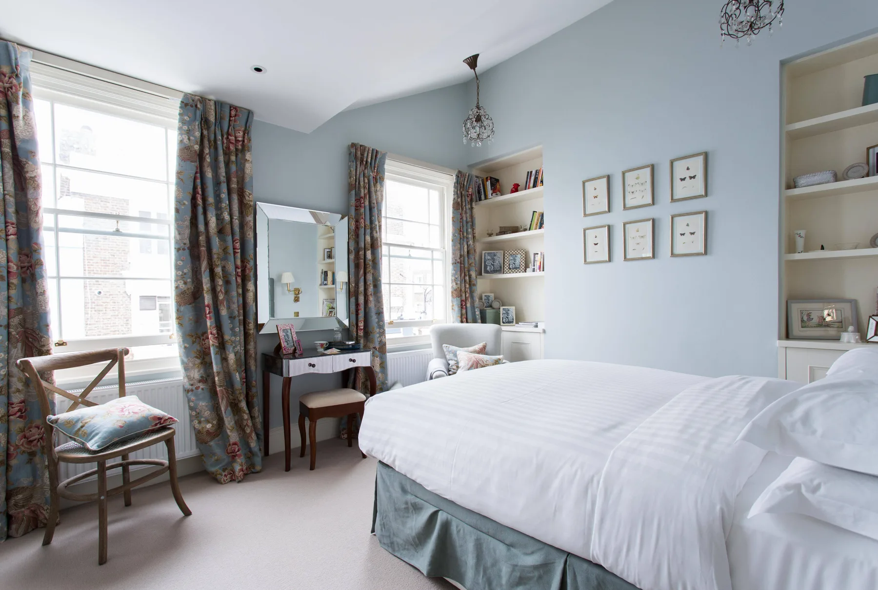 Un dormitorio luminoso con una cama, un escritorio pequeño y ventanas altas con cortinas.