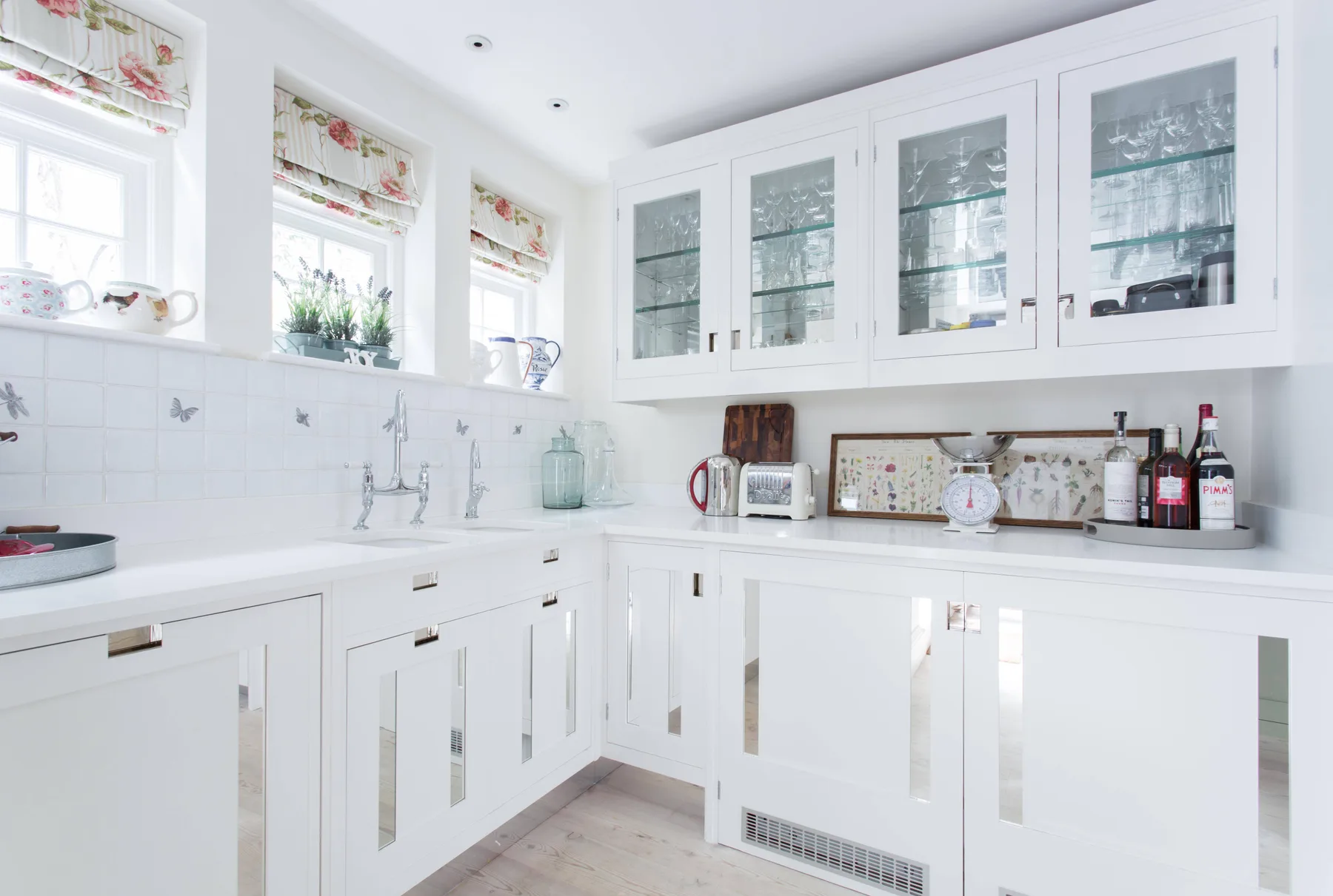 Una cocina blanca y luminosa con gabinetes de vidrio y un fregadero bajo las ventanas.