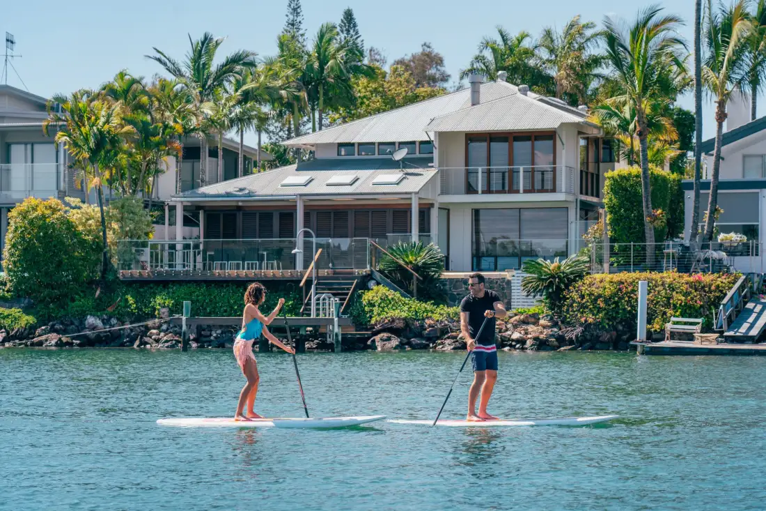 Varias personas practican paddle surf en un río frente a un edificio junto al agua.