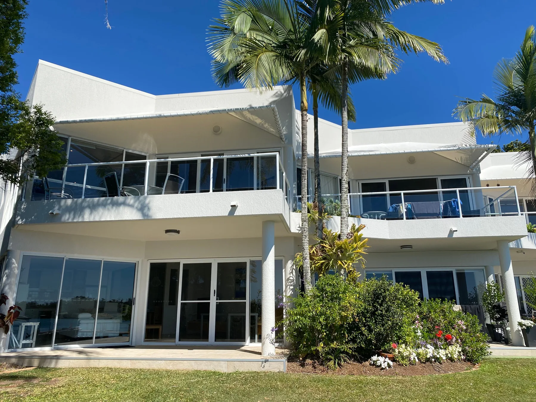 Edificio blanco de varias plantas con balcones, puertas de vidrio y jardín ajardinado.