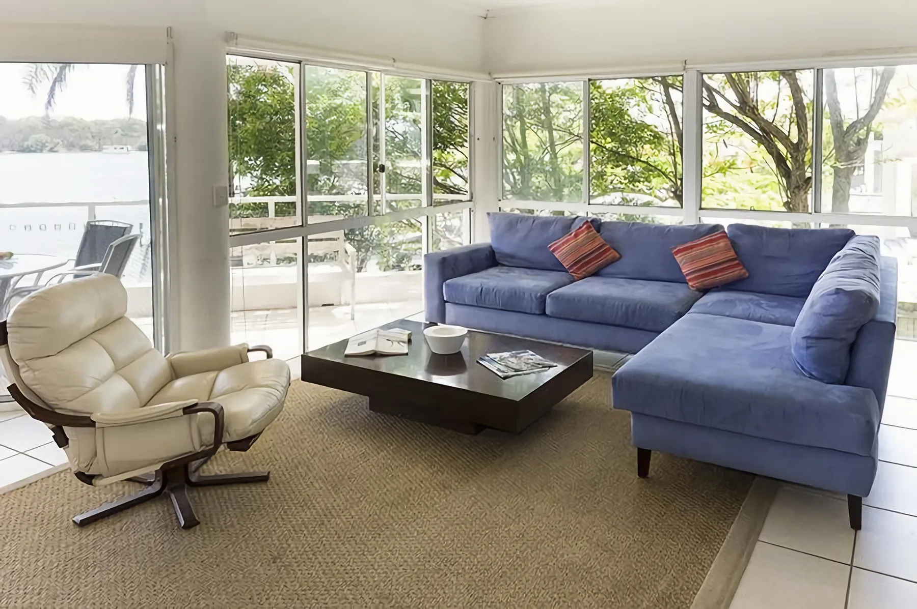 Una sala de estar con asientos azules y grandes ventanas con vistas a árboles y agua.
