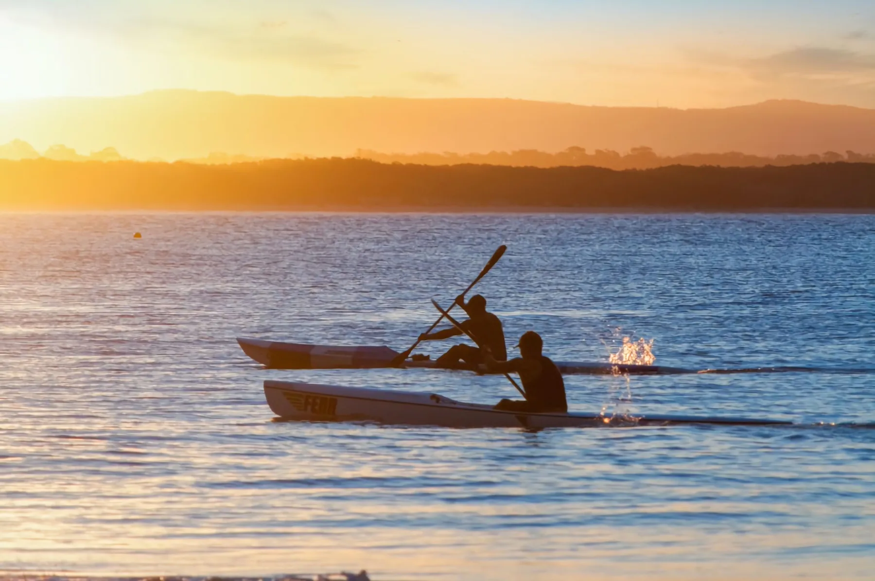 Dos personas reman en kayaks por un río tranquilo al atardecer.