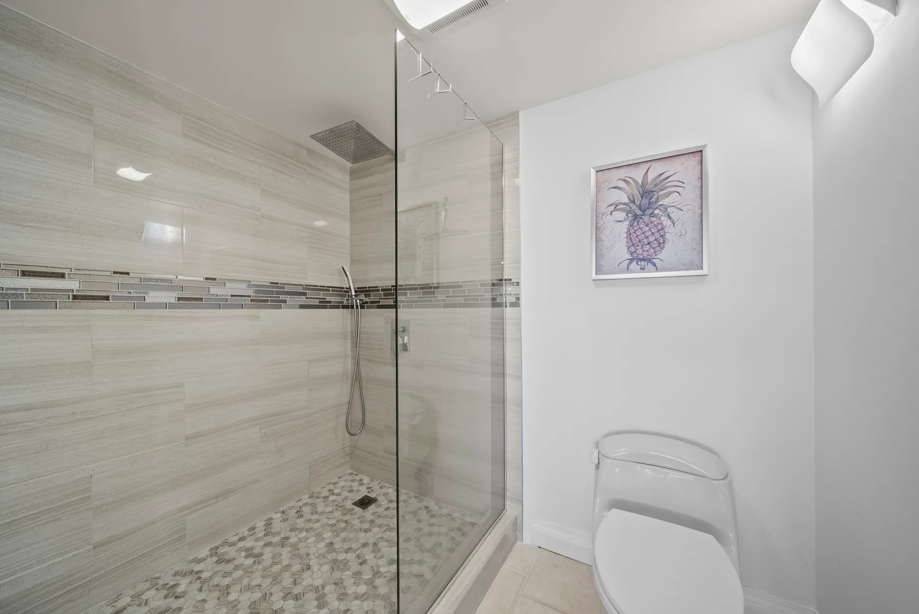 Un baño luminoso con ducha de vidrio y un cuadro enmarcado de una piña.