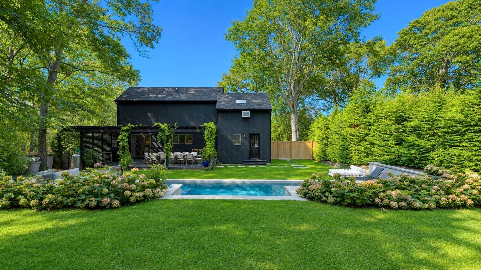 A dark modern house stands behind a swimming pool in a leafy garden.