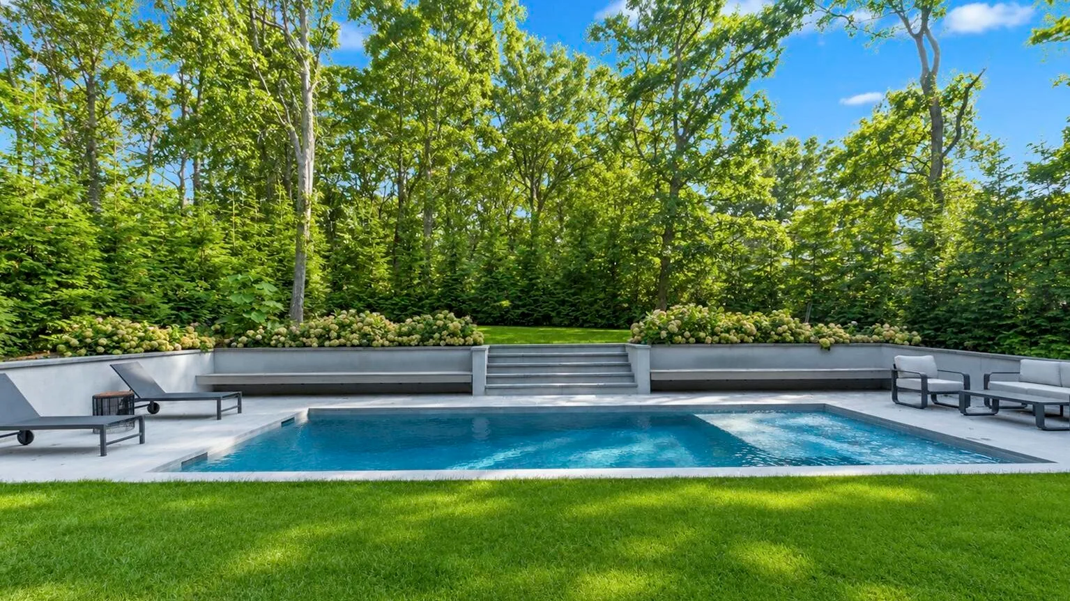 An outdoor swimming pool sits beside a lawn, with trees surrounding the patio.