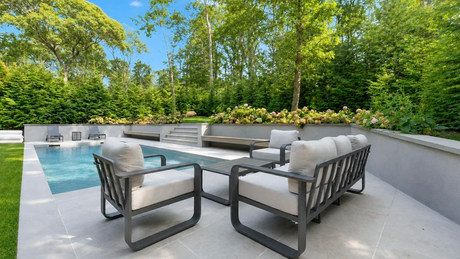 An outdoor patio with cushioned seating overlooks a swimming pool and leafy trees.
