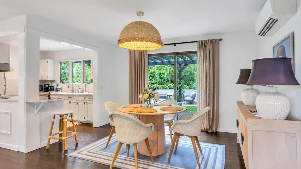 Dining area with a round table and chairs beside windows overlooking greenery.