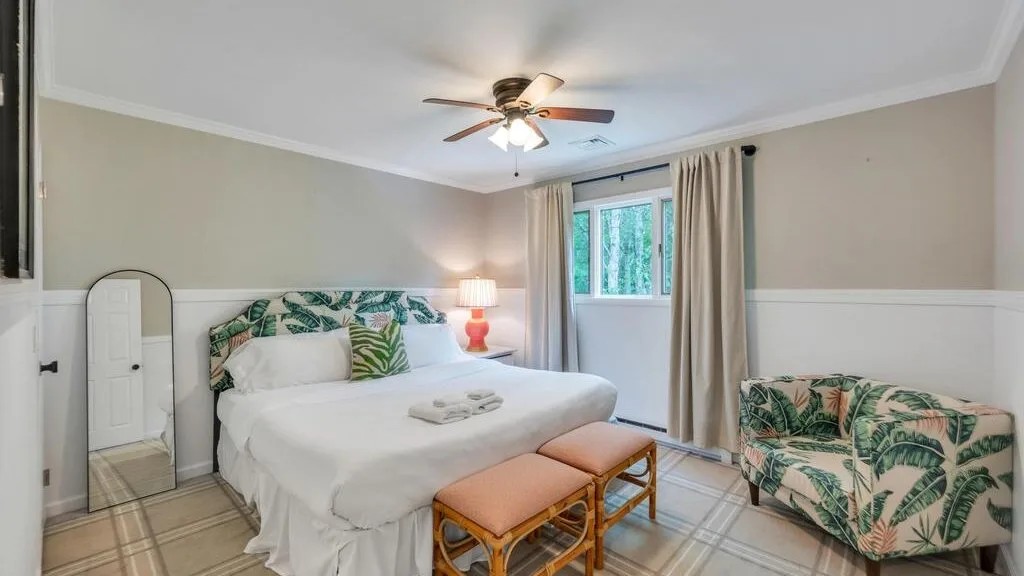 A bedroom with a white bed, ceiling fan, and tropical-patterned seating.