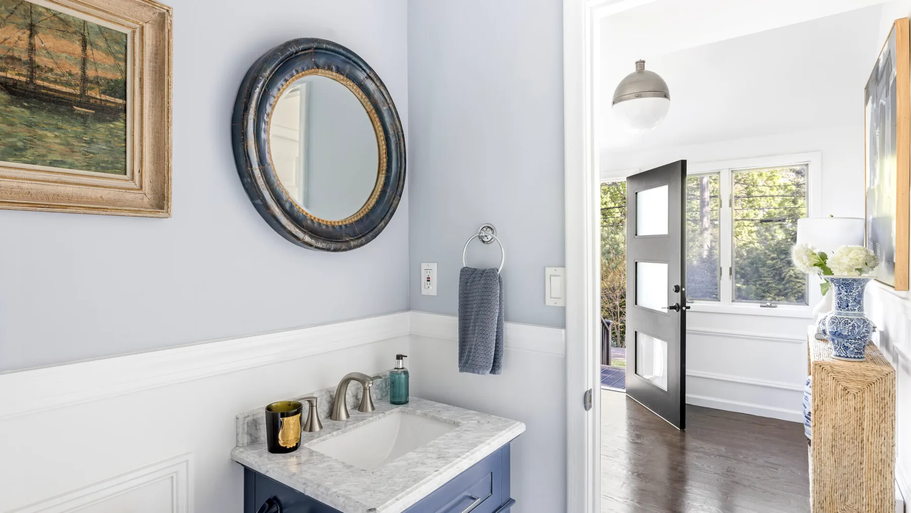 A bathroom vanity with a round mirror opens onto a bright adjoining room.
