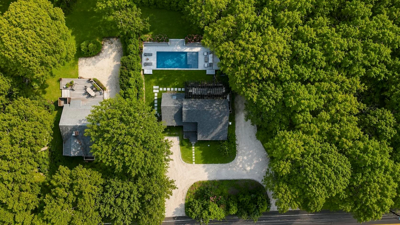 An aerial view shows a swimming pool, buildings, and a driveway among dense trees.