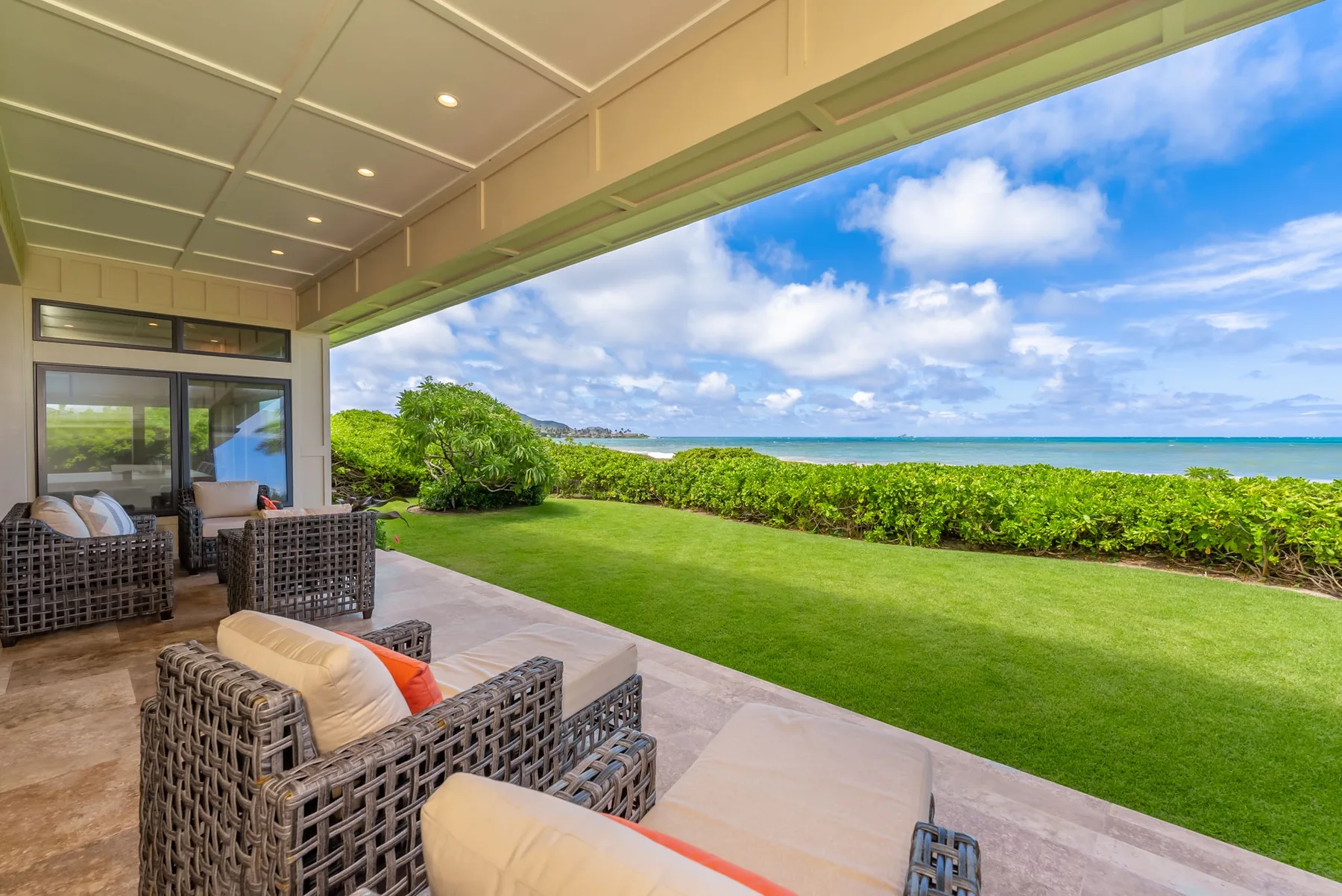 A covered patio with wicker seating overlooks a green lawn and ocean.