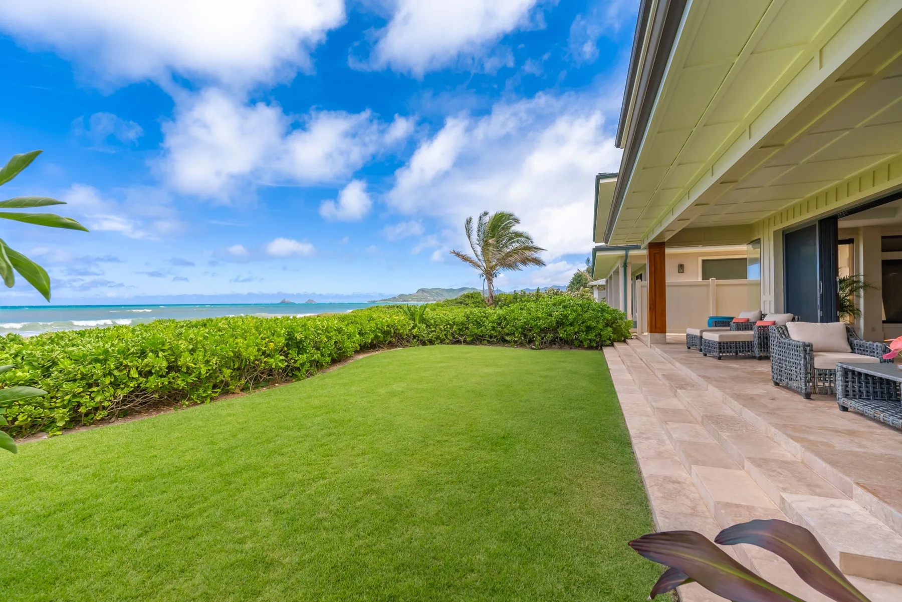 A grassy outdoor area beside a tiled patio, with ocean views beyond the hedge.